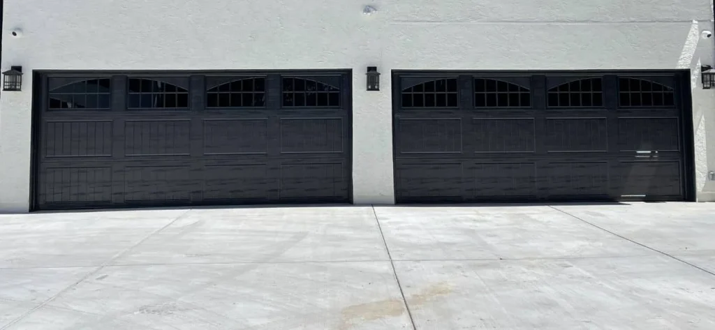 An exterior view of two modern, dark-paneled double garage doors with window inserts on a light stucco home, ideal for weighing steel vs wood for the environment.