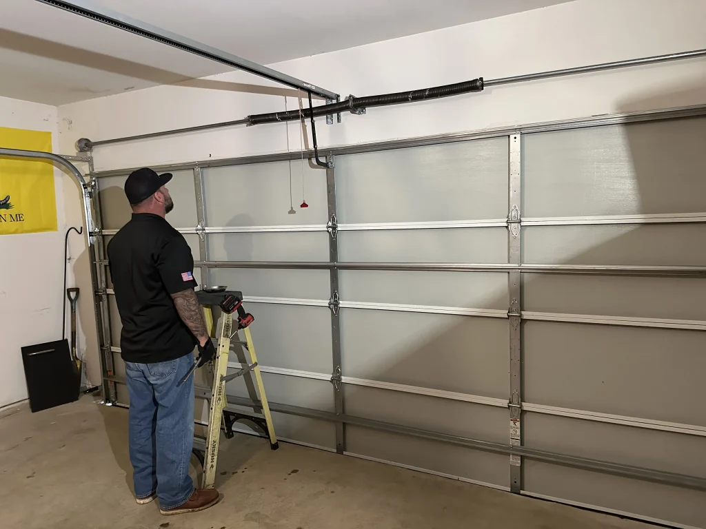 A technician standing next to a stepladder while inspecting the interior side of a multi-panel garage door system, ideal for discussing wood or steel garage door insulation options.