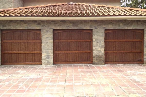 An exterior view of a three-car garage featuring elegant, warm-toned wooden doors embedded in a natural stone wall, perfect for tips on choosing the right garage door material.