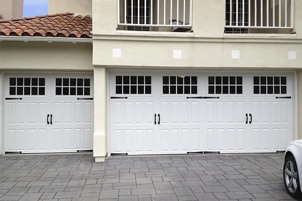 An exterior front view of a multi-car garage featuring classic white carriage-style doors with top windows and decorative black hardware, highlighting why garage door replacement matters for curb appeal.