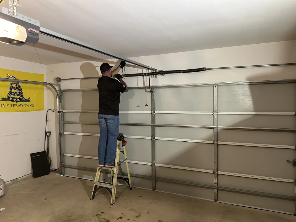 A technician standing on a stepladder inside a residential garage to repair the torsion spring and center bracket, demonstrating the signs you need emergency garage door repair in Celina, TX.